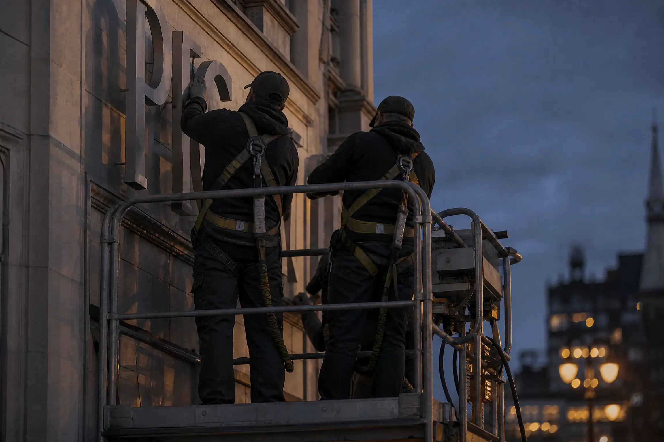Deux poseurs harnachés sur une nacelle, fixant des lettres découpées sur une façade à la tombée du jour